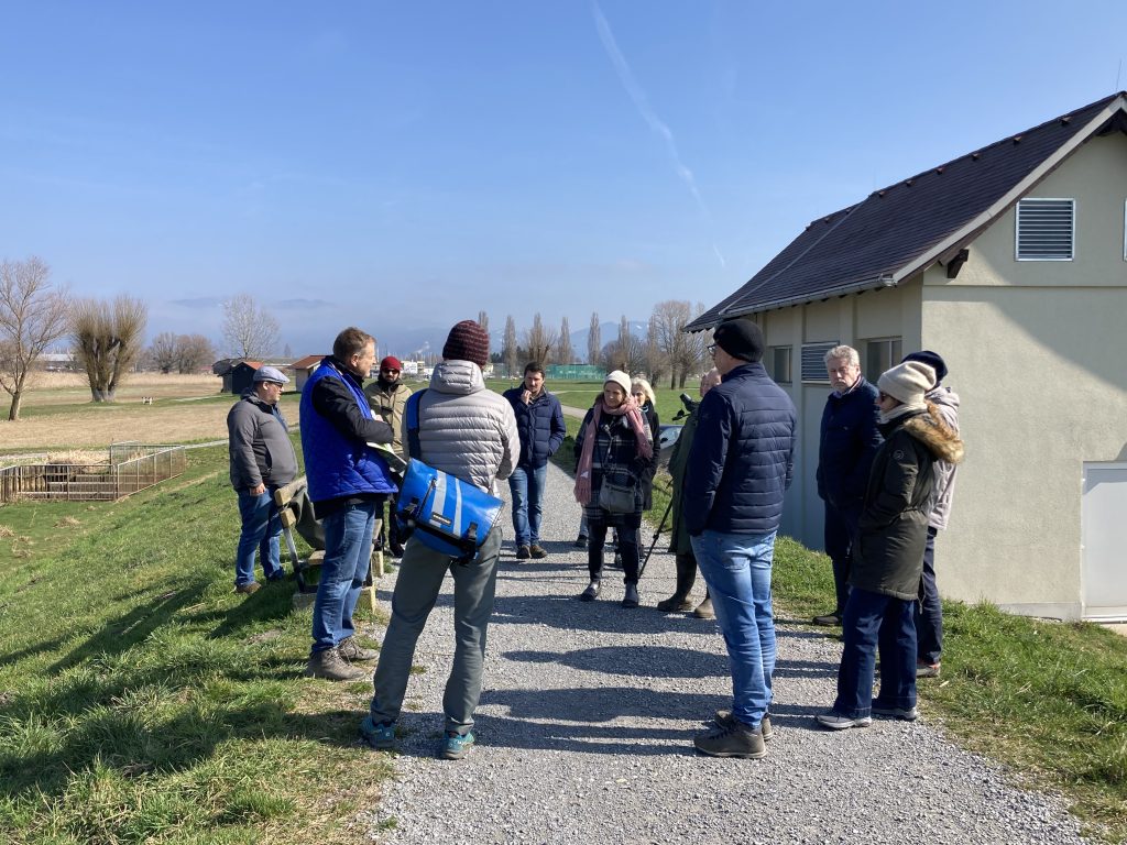 Das Bild zeigt eine Gruppe im Kreis aufgestellt auf einem Weg, umringt von grüner Vegetation. In der rechten Bildhälfte sieht man ein Haus.