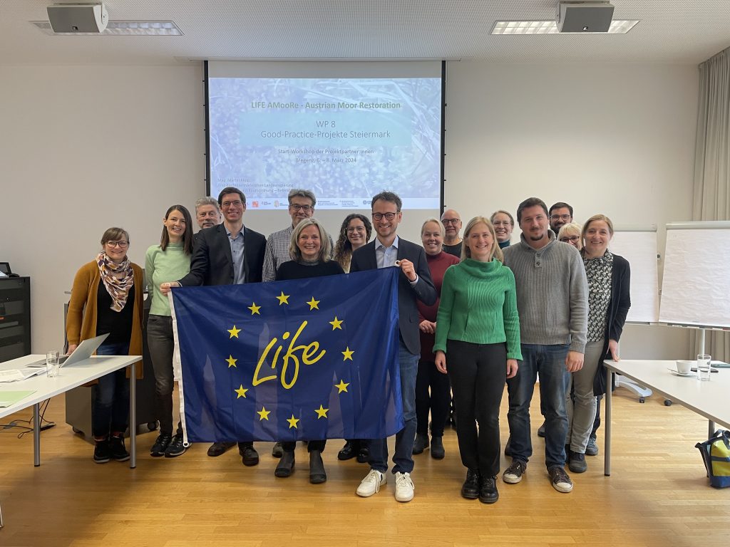 Das Bild zeigt eine Gruppe bestehend aus sechzehn Personen in einem geschlossenen Raum vor einer Leinwand. Zwei Personen halten eine große LIFE-Fahne in der Hand.