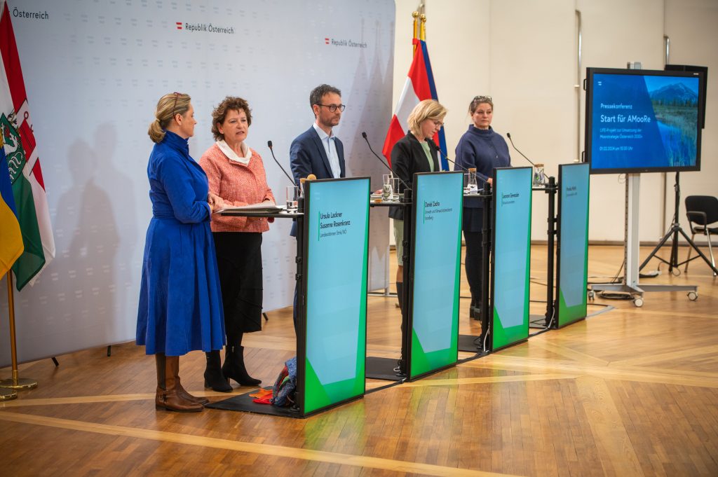 Das Bild zeigt fünf Personen stehend vor fünf Stehpulten vor einer Leinwand, umringt von Fahnen.